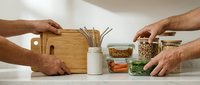 Male hands arranging wooden cutting board, ceramic utensil holder with cooking tools, and glass storage jars containing grains and dried foods on white kitchen counter surface.