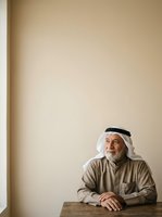 Senior Middle Eastern man wearing traditional white ghutra headdress and tan dishdasha robe, seated at wooden table against neutral beige wall. Suitable for cultural representation, diversity concepts, and portrait photography.