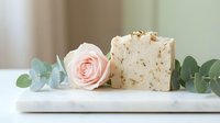 Handmade soap bar with oatmeal exfoliant next to fresh pink rose and eucalyptus leaves on white surface for natural skincare and wellness product marketing.