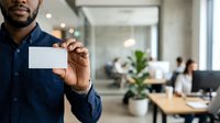 African American male professional in dark blue button-up shirt holding blank white business card in modern office environment with colleagues working at desks in background.