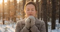 Young Caucasian woman in warm winter coat holding hot beverage mug outdoors during snowfall with peaceful expression and golden sunlight filtering through trees.