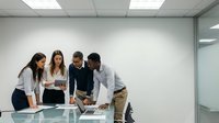 Diverse team of young professionals collaborating around table in modern office, reviewing digital tablet and documents during business meeting or project planning session.