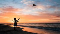 Young girl with long dark hair flying kite on sandy beach at sunset, silhouetted against golden sky and ocean waves, outdoor recreation and leisure activity.