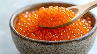 Fresh red caviar in ceramic bowl with wooden spoon, close up view of salmon roe pearls with bright orange color and glossy texture on light background.