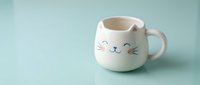 Adorable white ceramic cat mug with pink ears and cute facial expression on mint green background, kawaii style drinkware for coffee or tea lovers.