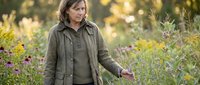 Middle-aged Caucasian woman gardener in olive green jacket tending to autumn flower garden with purple blooms and golden foliage during sunset hour.