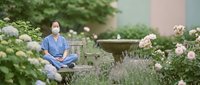 Young Asian woman in blue medical scrubs relaxing on wooden bench in peaceful hospital garden surrounded by blooming flowers and greenery.