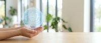 Hand holding glowing digital brain hologram over wooden desk in modern office interior with plants and natural light for AI technology concepts.