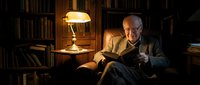Elderly Caucasian man reading book in cozy library with warm lamp light, surrounded by bookshelves. Perfect for literature, education, retirement lifestyle concepts.