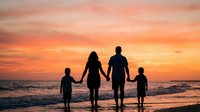 Family of four holding hands watching sunset on beach with orange sky and ocean waves creating peaceful silhouette moment for vacation memories.