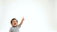 Young toddler boy with dark curly hair, appearing excited and pointing upward with enthusiasm against white background for advertising and commercial use.