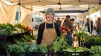 Young Caucasian female farmer in beige apron and green beanie smiling at outdoor farmers market stand with fresh organic vegetables and leafy greens for healthy eating.
