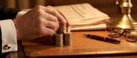 Businessman hand stacking coins on wooden desk with documents and scales, representing financial planning, investment growth and wealth management concepts.
