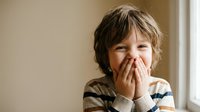 Young boy with tousled hair covering mouth with hands while laughing in striped sweater at home showing playful childhood emotions and joy.