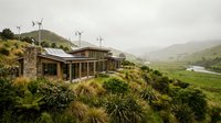 Modern eco-friendly house with wind turbines on hillside overlooking green valley. Sustainable architecture blends with natural landscape in rural setting.