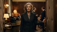 Professional mature Caucasian businesswoman in elegant dark suit standing confidently in upscale hotel lobby with warm lighting and business people in background.