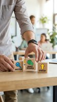 Male professional in casual business attire arranging wooden blocks with analytics, market research, and strategy icons in modern office environment with colleagues and plants in background.