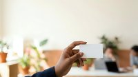 Man with dark skin tone holding blank white business card in office environment with colleagues and potted plants in background.
