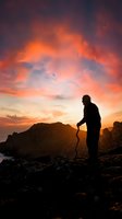 Silhouette of male hiker with walking stick standing on rocky mountain summit against dramatic orange sunset sky for adventure and outdoor recreation concepts.