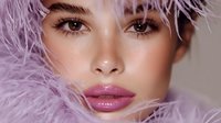 Close up portrait with pink feathers framing face, highlighting glossy mauve lips, defined brows and luminous skin against soft neutral background.