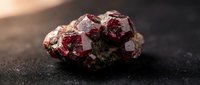 Raw red garnet crystal cluster with deep burgundy faceted gemstones on dark surface, natural mineral specimen for jewelry making and geological collection.