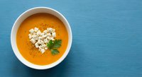 Creamy orange soup with white cheese crumbles and fresh herbs in white bowl on blue wooden background. Comfort food for restaurant menu design.