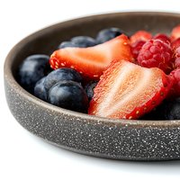Fresh mixed berries including sliced strawberries, whole blueberries and raspberries served in modern speckled gray ceramic bowl, close-up view on white background.