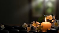 Spa candle glowing softly surrounded by plumeria flowers and smooth black stones creating peaceful zen atmosphere for relaxation and wellness treatments.