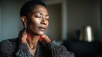 African woman with neck pain, massaging sore area with pained expression. Emotional portrait showing discomfort and stress in soft natural lighting.