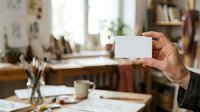 Hand holding blank white card in creative studio workspace with desk, supplies, and natural light.