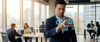 Young male professional of Caucasian ethnicity in business attire interacting with digital holographic data visualization in modern office environment with colleagues working in background.