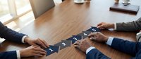 Business team assembling puzzle pieces on wooden conference table representing teamwork, collaboration and problem-solving in corporate meeting environment.
