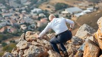 Man climbing rocky mountain terrain overlooking suburban landscape with houses and hills in background during golden hour light.