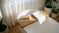 Wooden bath caddy with white towel and candle on bathtub edge, minimalist bathroom decor with natural light and potted plants for spa relaxation.