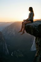 Young woman meditating on cliff edge at sunset overlooking mountain valley. Peaceful mindfulness moment in nature for wellness and spiritual growth concepts.