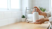 Woman with medium skin tone dispensing liquid soap from a white pump bottle in a bright, minimalist bathroom setting with natural light for hygiene and wellness content.