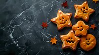 Star-shaped and round orange Halloween cookies with spooky faces on dark marble background with decorative autumn leaves, festive holiday baking.