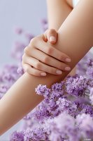 Elegant female hands with natural manicure resting on arm surrounded by fresh lavender flowers against soft purple background.