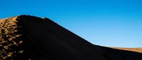 Young adult male with light skin tone, wearing casual outdoor attire, climbing a steep volcanic sand dune under clear blue sky. Suitable for travel, adventure, fitness and lifestyle content.