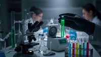 Female scientist in black gloves holding green test tube in modern laboratory with microscope and colorful samples for research analysis.