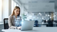 Young professional businesswoman working on laptop in modern office with digital network connections and data visualization graphics overlay.