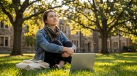 Young Asian woman student sitting on grass with laptop computer in autumn university campus park, looking up thoughtfully during outdoor study session.