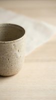 Handmade ceramic cup with speckled glaze texture sitting on light wooden surface with blurred neutral background, suitable for home decor and lifestyle content.