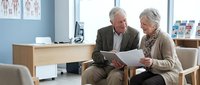 Senior Caucasian couple reviewing medical documents together in modern healthcare office waiting room with anatomical charts on wall.
