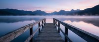 Wooden pier extending into misty lake at dawn with mountain silhouettes and pink purple sky reflection creating peaceful morning atmosphere for meditation retreat.