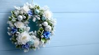 Festive winter wreath with white roses, blue hydrangeas, and silver ornaments hanging on light blue wooden wall for holiday home decoration.