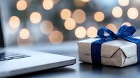 Silver wrapped present with navy blue ribbon bow next to laptop on desk with soft glowing bokeh lights creating festive holiday atmosphere.
