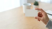 Person of medium skin tone holding blank white business card at wooden desk with coffee mug and plant, suitable for branding and corporate presentations.