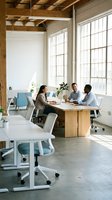 Diverse team of professionals collaborating at wooden table in modern loft office with industrial ceiling, large windows, and contemporary furniture for corporate workspace design.