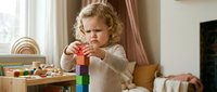 Young blonde toddler boy playing with colorful wooden building blocks in bright playroom, developing motor skills and creativity through educational toys.